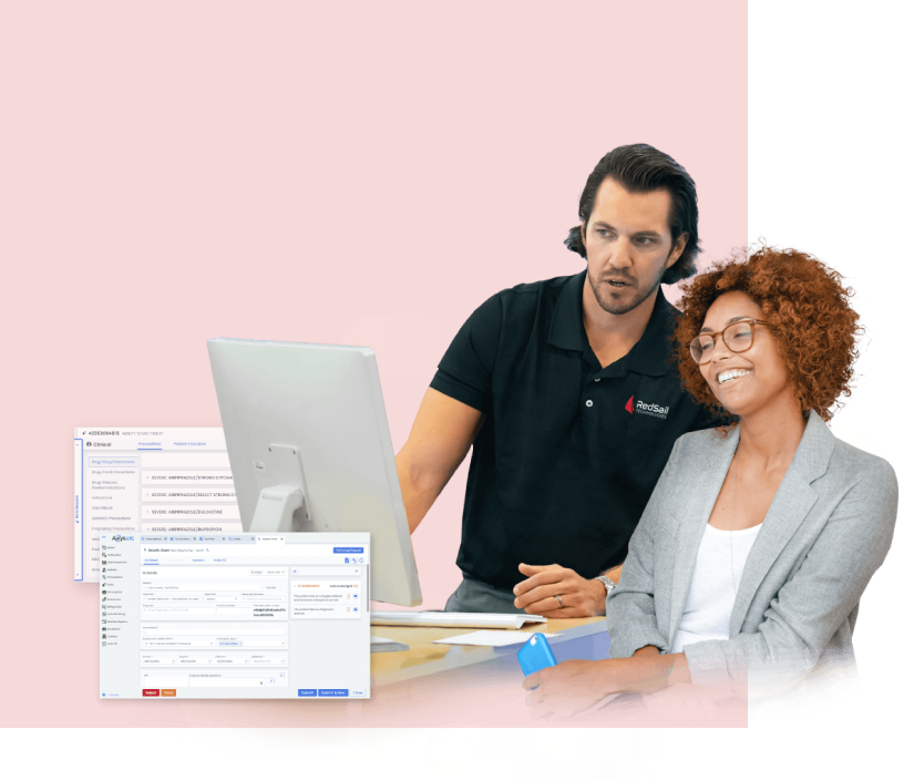 Two individuals are reviewing pharmaceutical software on a computer screen. The man, dressed in a dark polo shirt bearing the company logo, stands and gestures toward the display. The woman, seated and wearing a gray blazer and glasses, gazes at the screen with a pleased expression. Computer screenshots are displayed in the foreground.