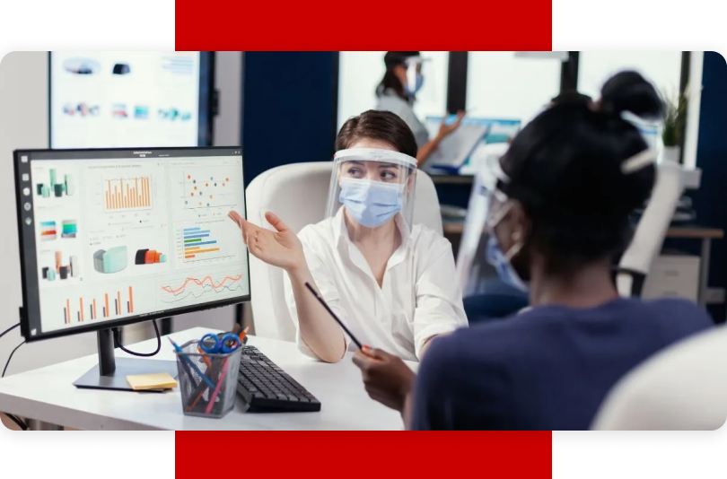 Office environment showing a Caucasian woman wearing a face shield and medical mask, gesturing towards a computer screen that displays multiple charts and graphs. The computer is centered. An African American woman with her back to the viewer is listening intently to the Caucasian woman. Both women are professionally dressed in white and blue shirts respectively. Another office worker wearing a face mask is in the background. This conveys data analysis in the cosmetic industry facilitated by enterprise resource planning (ERP) systems.