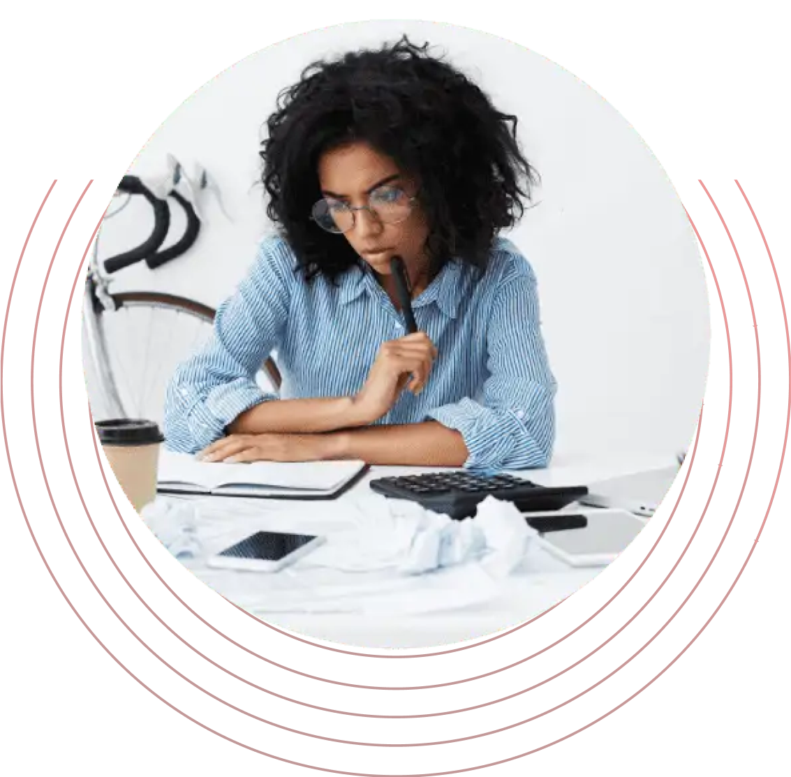 A woman sitting at a desk with a pen in her mouth, looking thoughtfully as she contemplates her work. She is wearing casual clothing and has a focused expression on her face. The setting includes some art supplies around her.