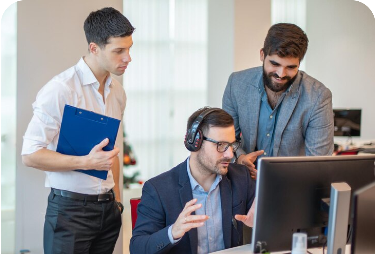 A group of men gathered around a laptop, intently looking at the computer screen. They are indoors and appear to be engaged in work-related activities. The setting includes a wall behind them, and the men are dressed in various clothing styles.